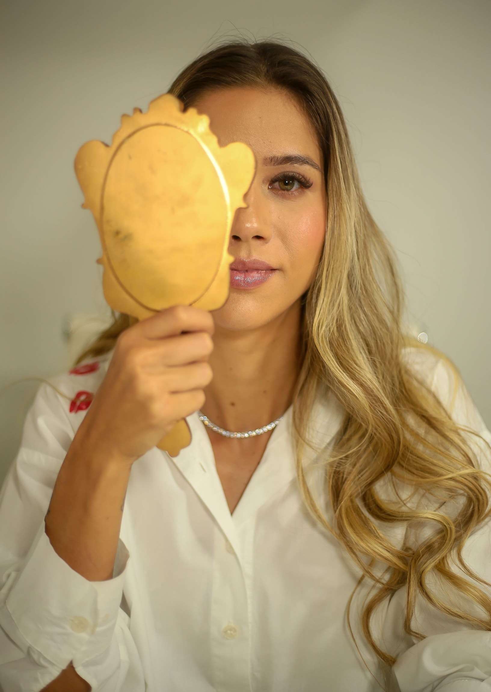 Dra. Alice Fortes segurando um espelho de mão dourado que cobre metade do rosto.